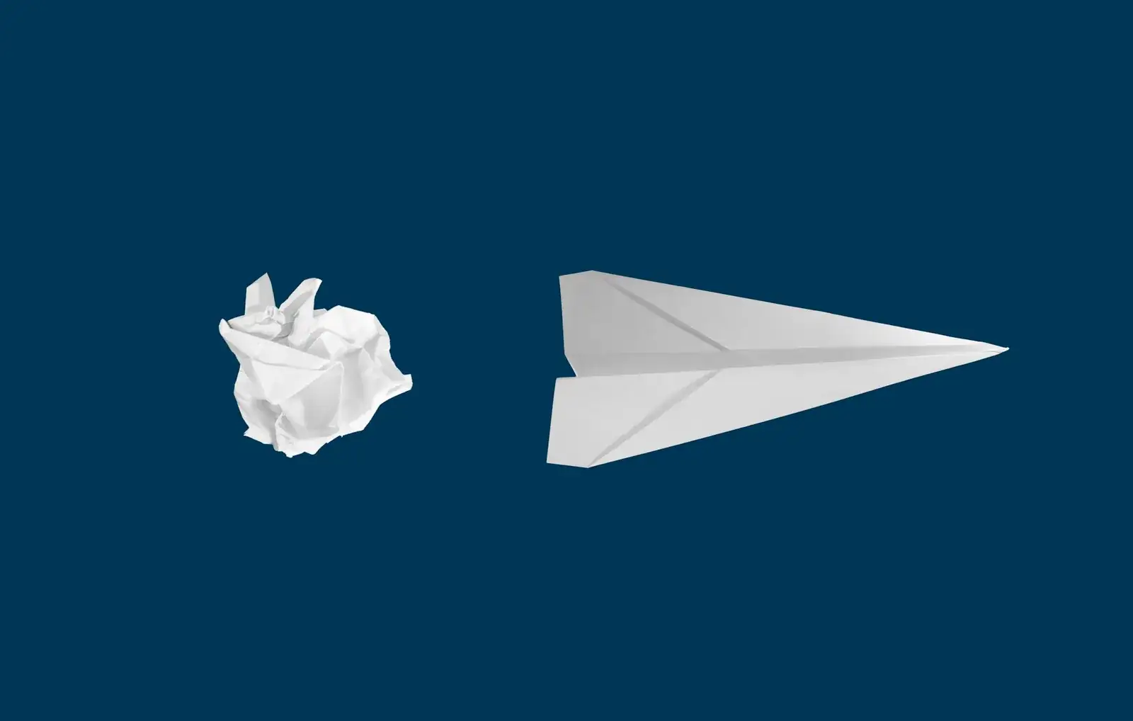 Avion en papier à côté d’une boule de papier froissé, sur un fond bleu nuit